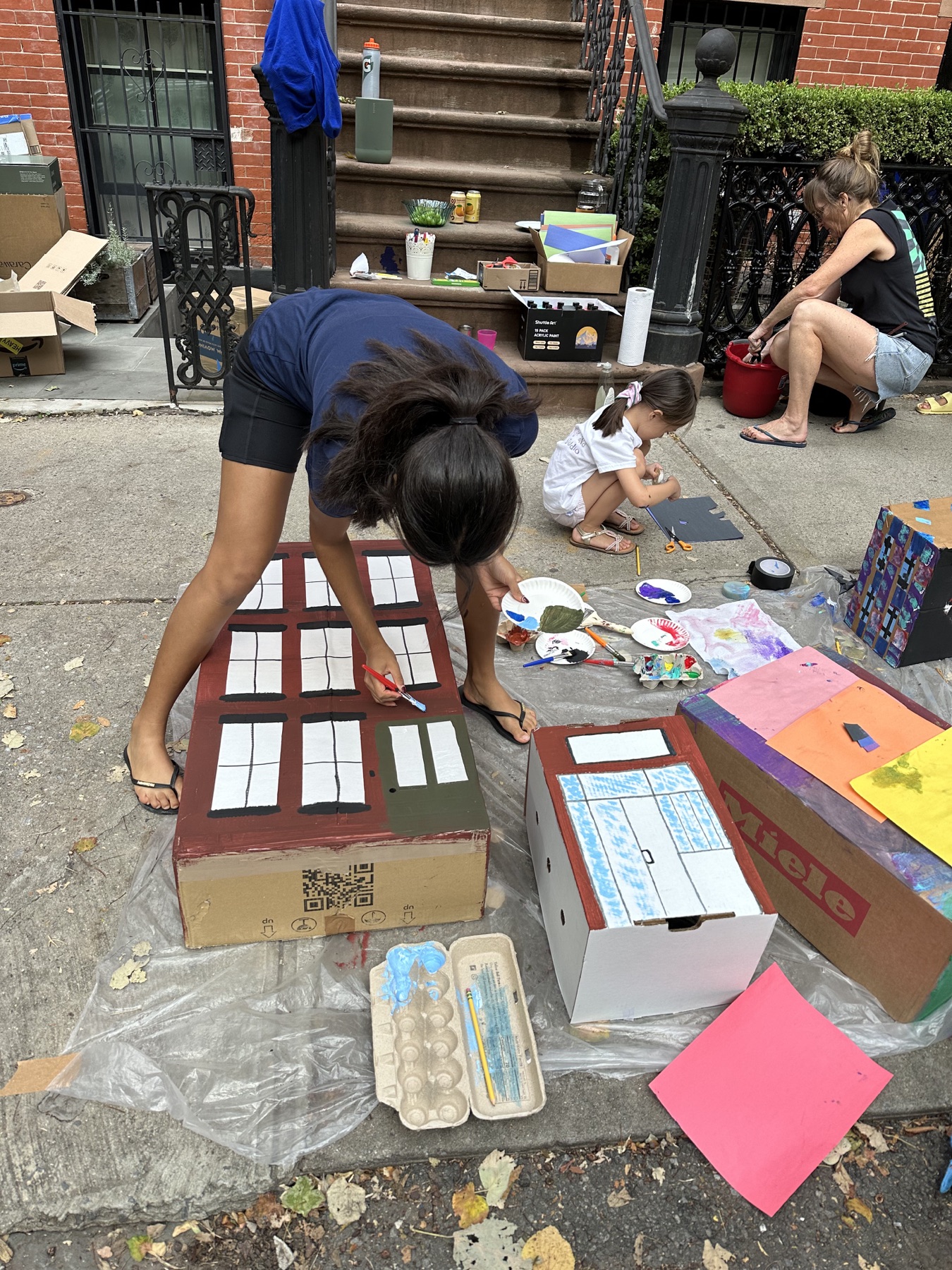 Neighbors painting a group of colorful cardboard buildings outside a brownstone
